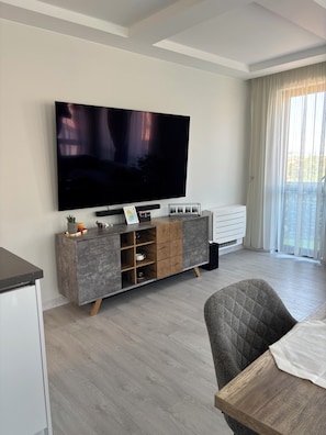 Un saló modern amb televisió de pantalla plana, un moble de pino amb calaixos, una taula de menjador i una finestra gran amb cortines lleugeres.