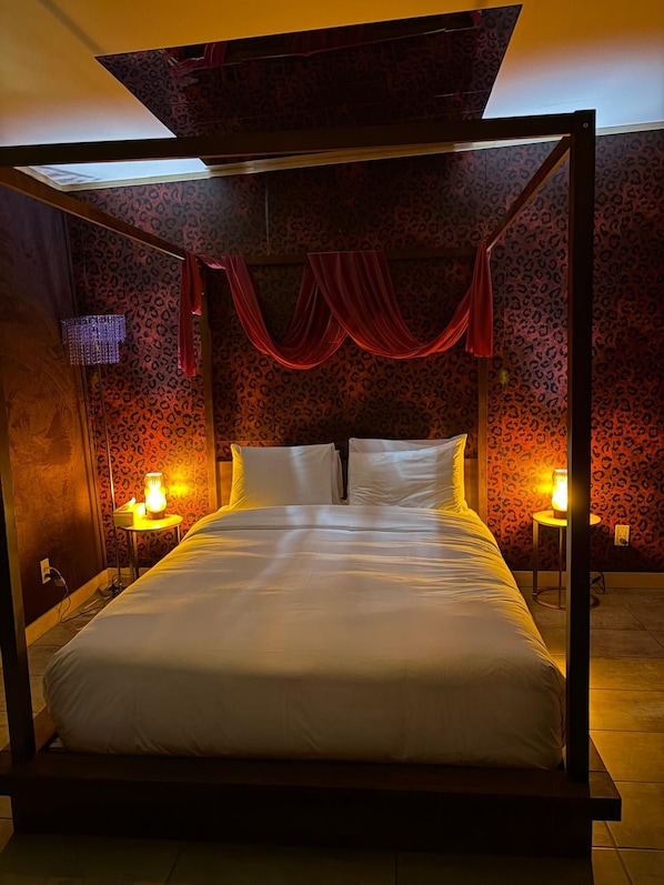 A four-poster bed with white linens, two bedside tables with lamps, and a leopard print wallpaper.