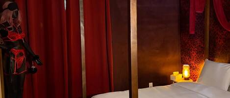 A four-poster bed with white bedding, a nightstand with a lamp, and a red curtain.