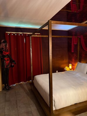 A four-poster bed with white bedding, a nightstand with a lamp, and a red curtain.