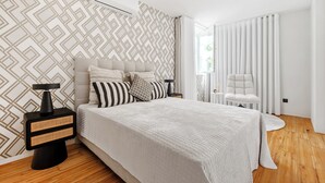 Un dormitorio moderno con un papel tapiz de diseño geométrico, una cama con varios almohadones, una mesita de noche con lámpara y una ventana con cortinas.