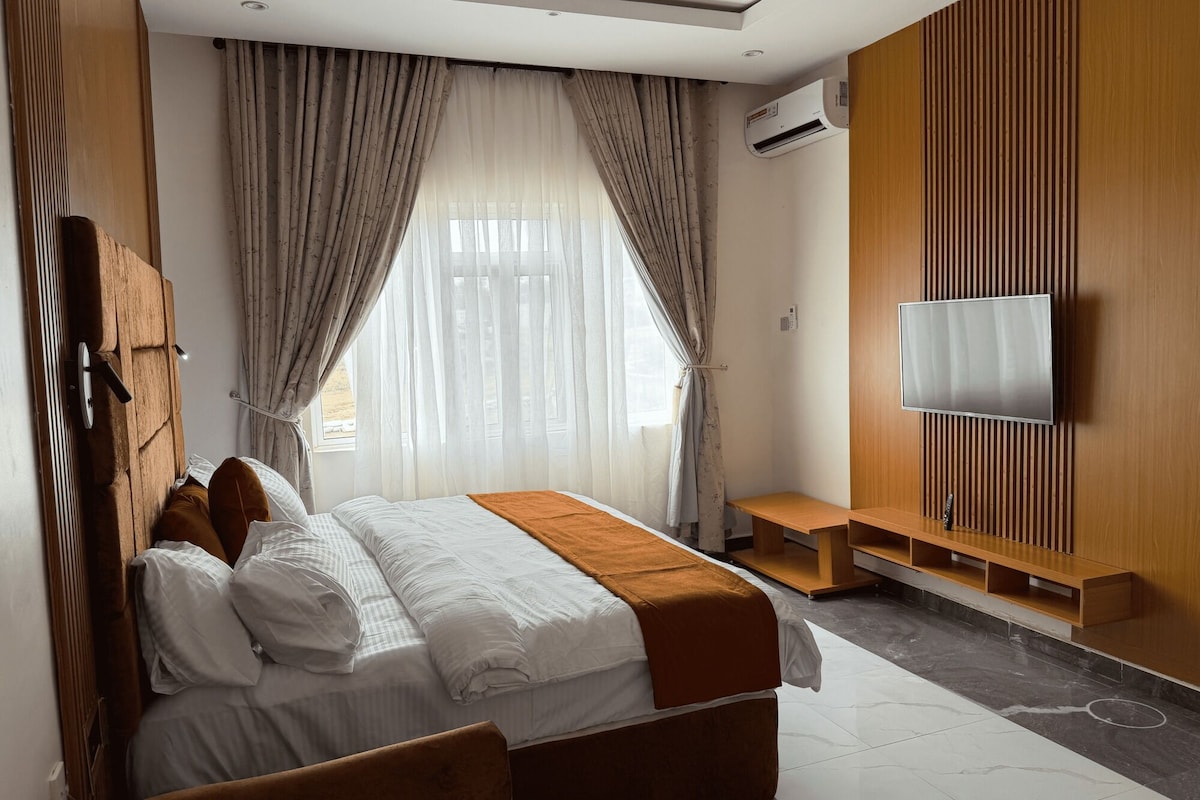 Una habitación de hotel moderna con cama, cortinas, televisor y pared de paneles de madera.