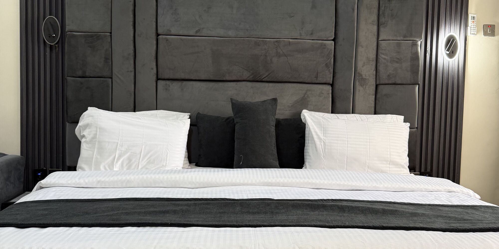 Una cama con cabecera oscura, almohadas blancas y oscuras, y una manta negra.