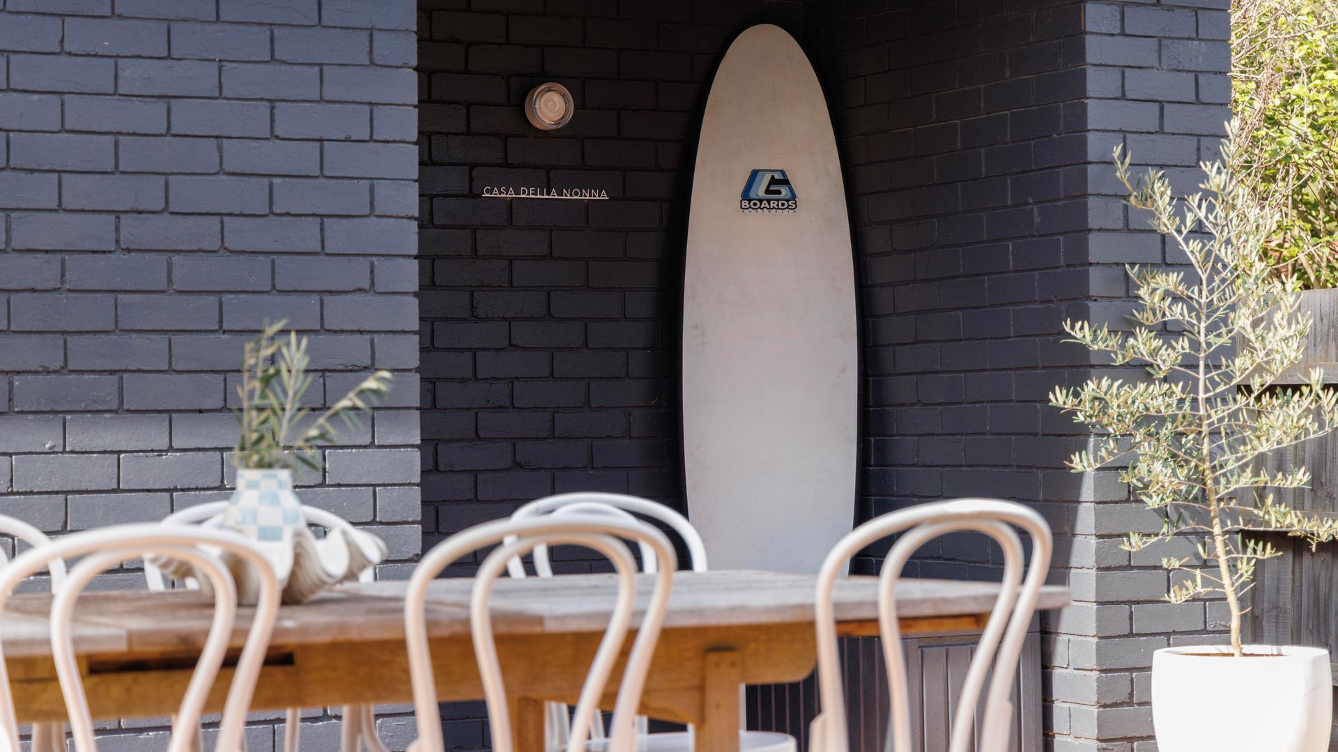 A surfboard leaning against a brick wall with a logo, a wooden table with chairs, and a potted plant.