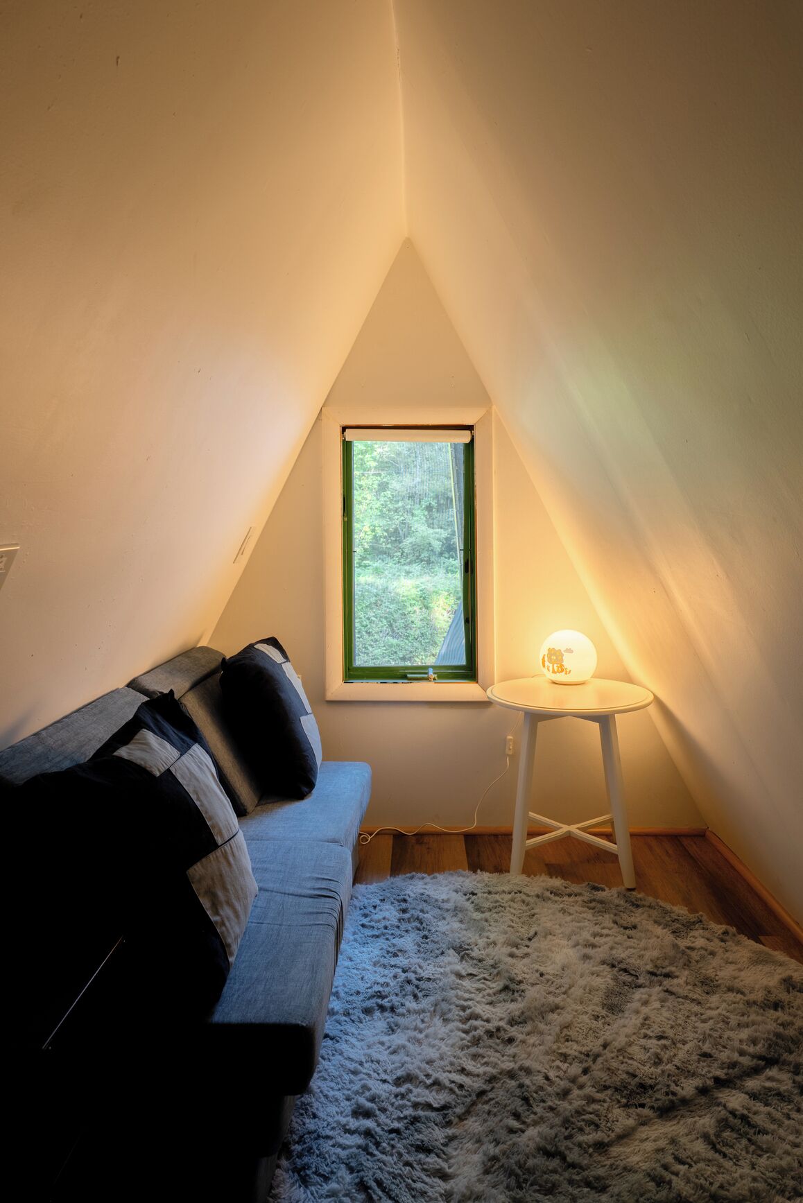 A cozy attic room with a sloped ceiling, a window with a view of greenery, a small table with a lamp, and a sofa with a fluffy rug.