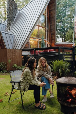 Two individuals seated on a bench outdoors, with a fire pit and a wooden structure in the background. - Fern Haus  Hiking, Hot Tub & River Access (Gold Bar)