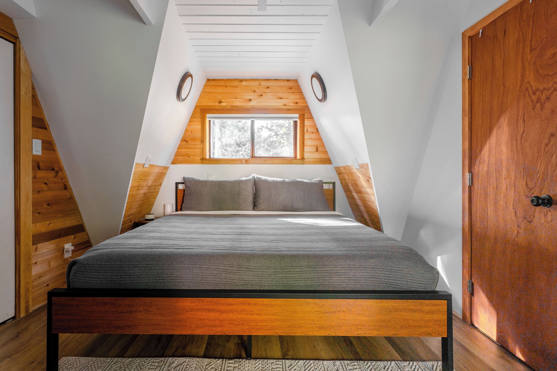 A bedroom with a sloped ceiling, a bed with a wooden frame, a window, and a door.