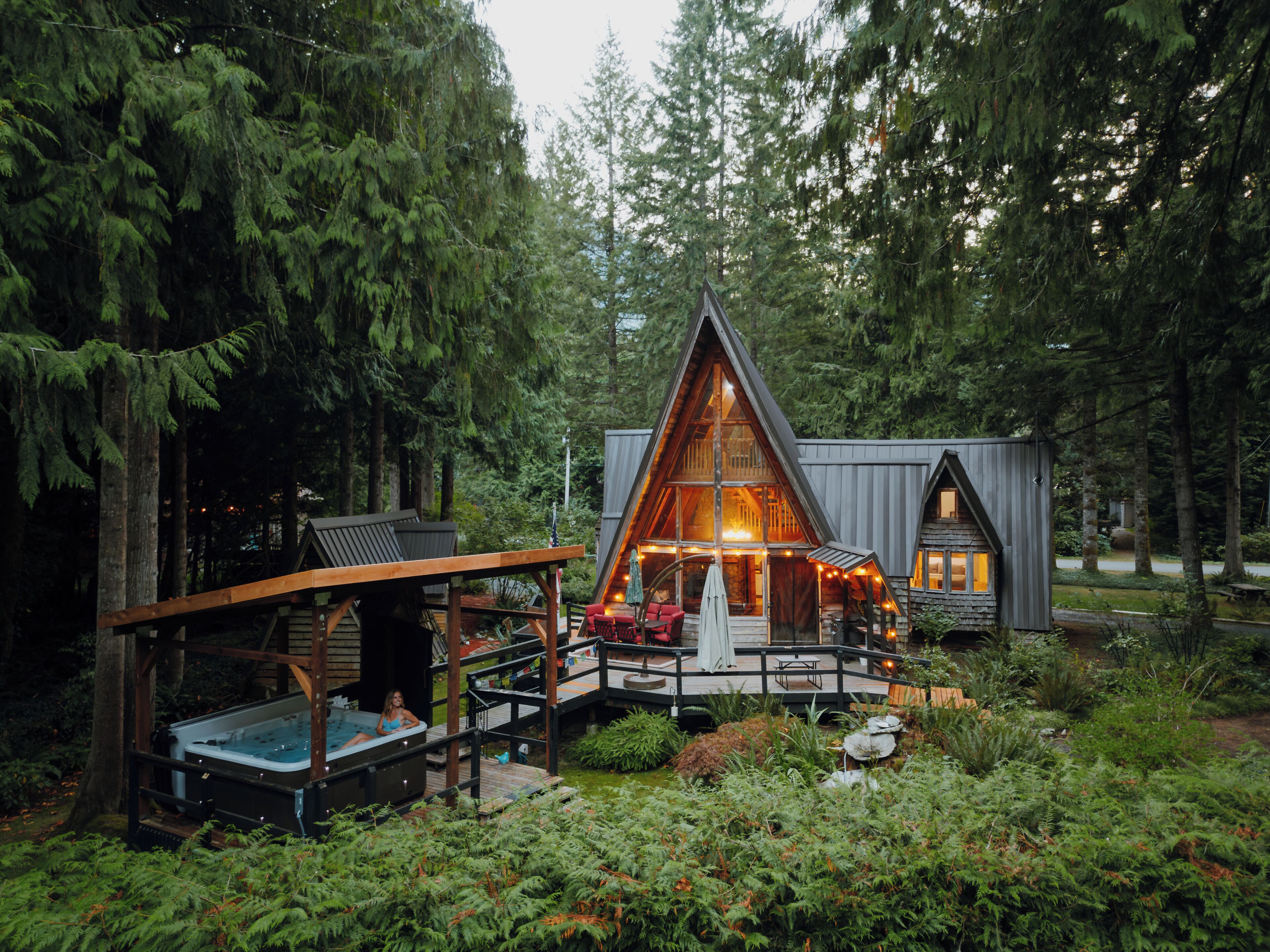 A cabin with a hot tub, deck, and outdoor seating area surrounded by dense forest.
