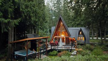 A cabin with a hot tub, deck, and outdoor seating area surrounded by dense forest.