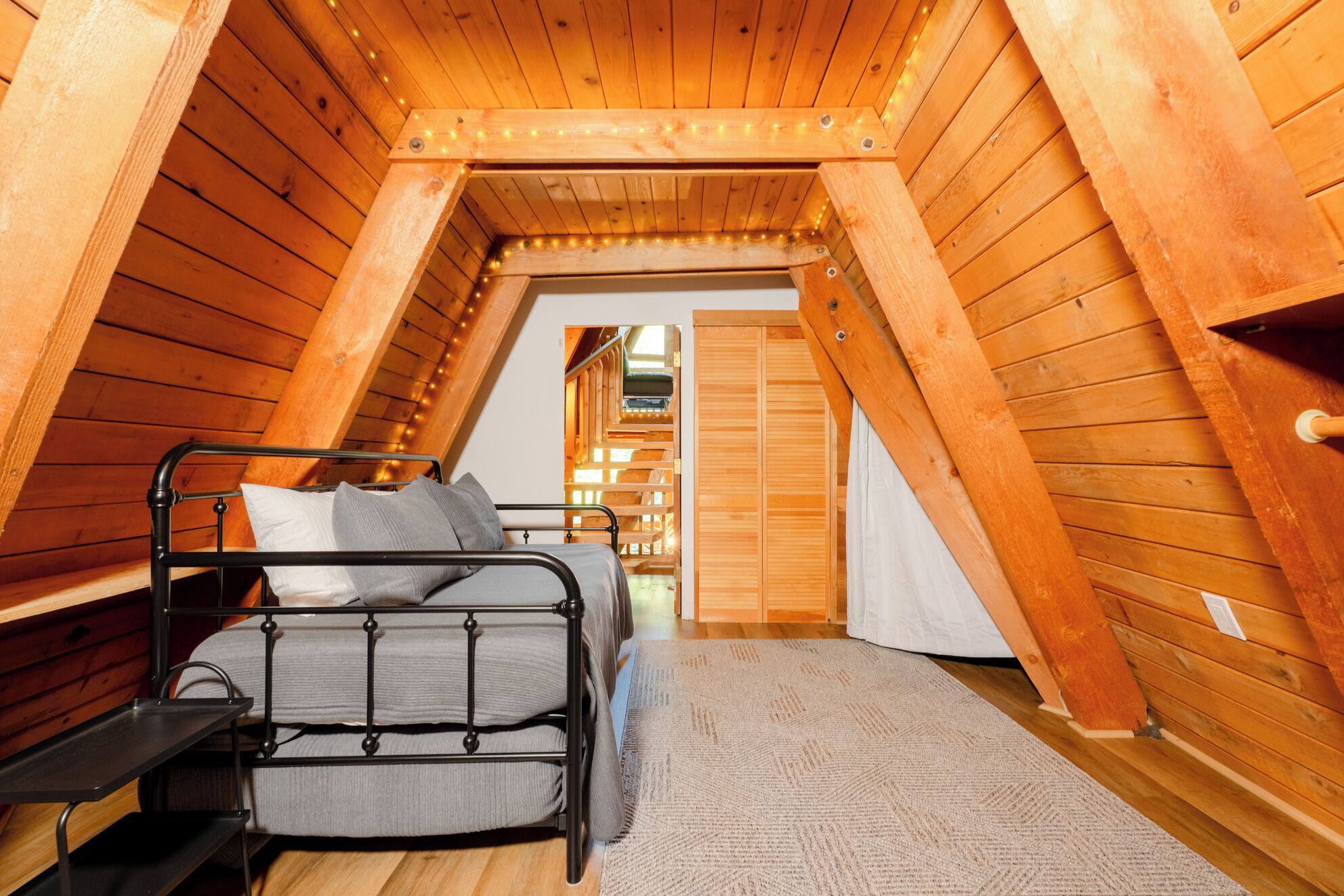 A small, wooden attic bedroom with a metal bed frame, a bench, and a view of a wooden hallway.