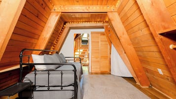 A small, wooden attic bedroom with a metal bed frame, a bench, and a view of a wooden hallway.