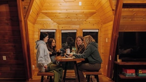 Eine Gruppe Personen sitzt an einem Holztisch in einer Hütte mit schrägem Dach und einer Pendelleuchte.