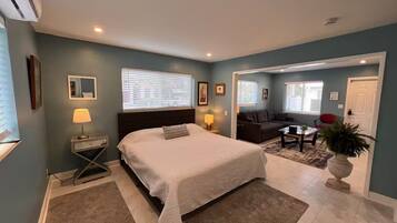 A bedroom with a bed, bedside lamps, a window with blinds, a nightstand, and a door leading to a living area with a sofa and a coffee table.