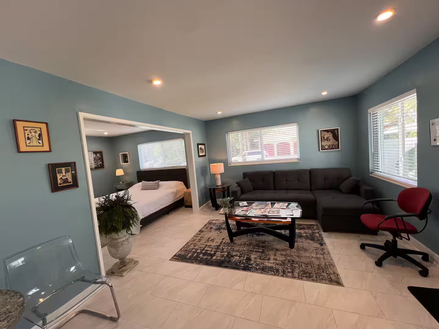 A room with a bed, a sofa, a coffee table, and a chair. There are windows with blinds, a lamp, and framed pictures on the walls.