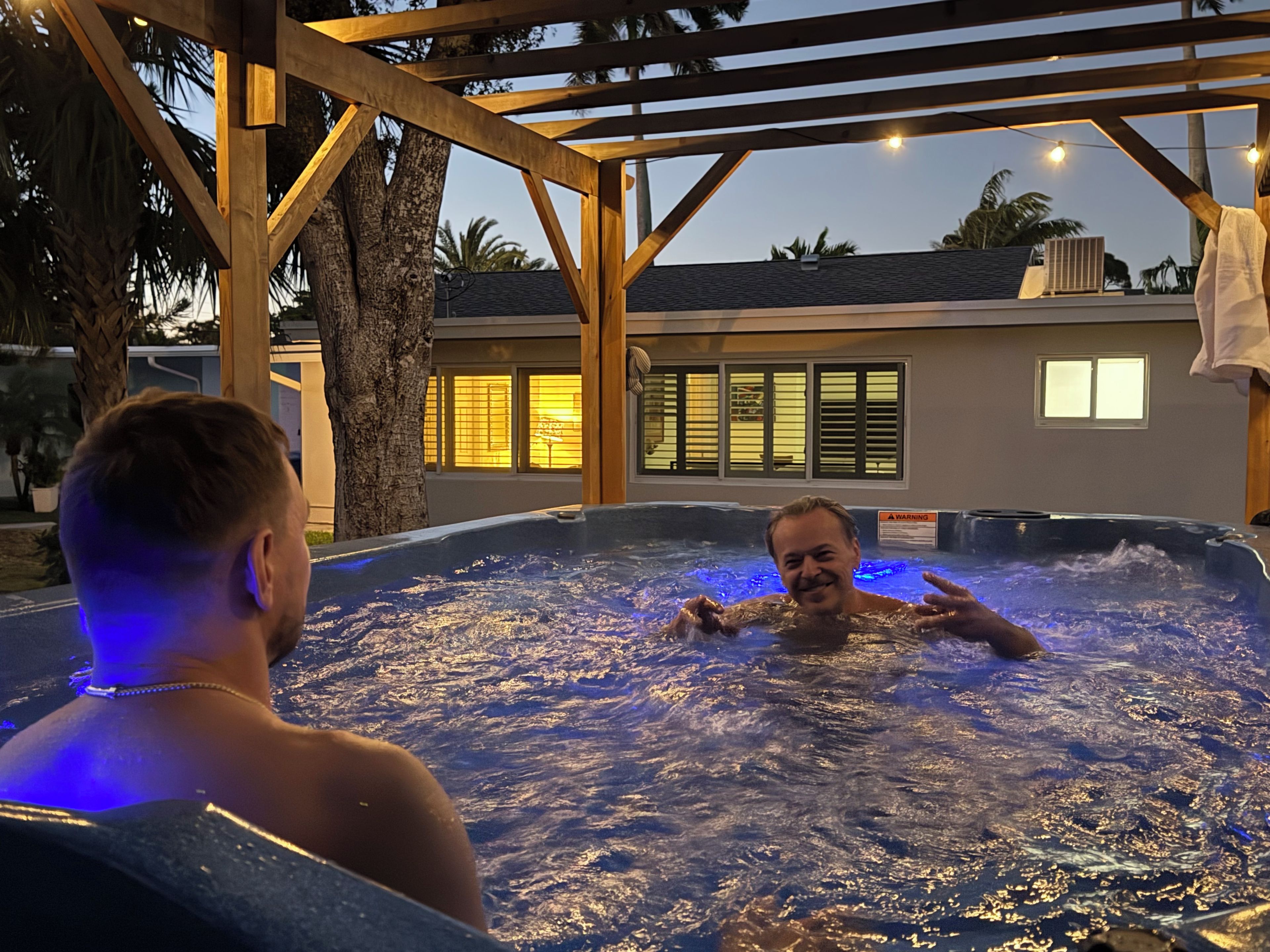A hot tub with a wooden pergola overhead, surrounded by palm trees and a building with lit windows in the background.