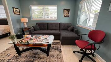 A living room with a sectional sofa, a red office chair, a glass coffee table with magazines, a lamp, and a window with blinds.