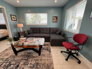 A living room with a sectional sofa, a red office chair, a glass coffee table with magazines, a lamp, and a window with blinds. - The Pines Hideaway 508, One bedroom studio suite (Wilton Manors)