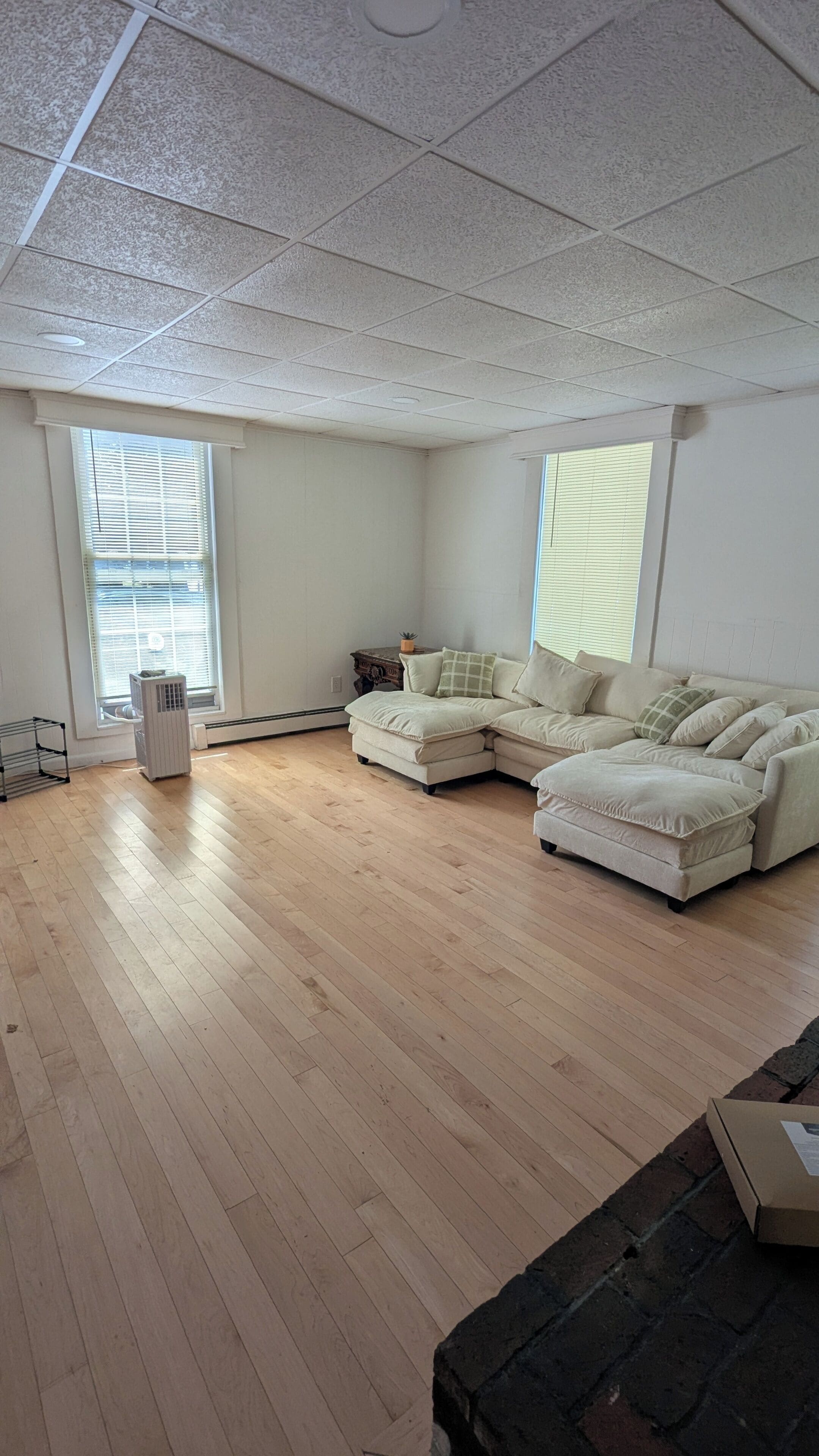 A spacious living room with a sectional sofa, a standing heater, and a window with blinds.