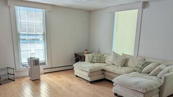 A spacious living room with a sectional sofa, a standing heater, and a window with blinds.