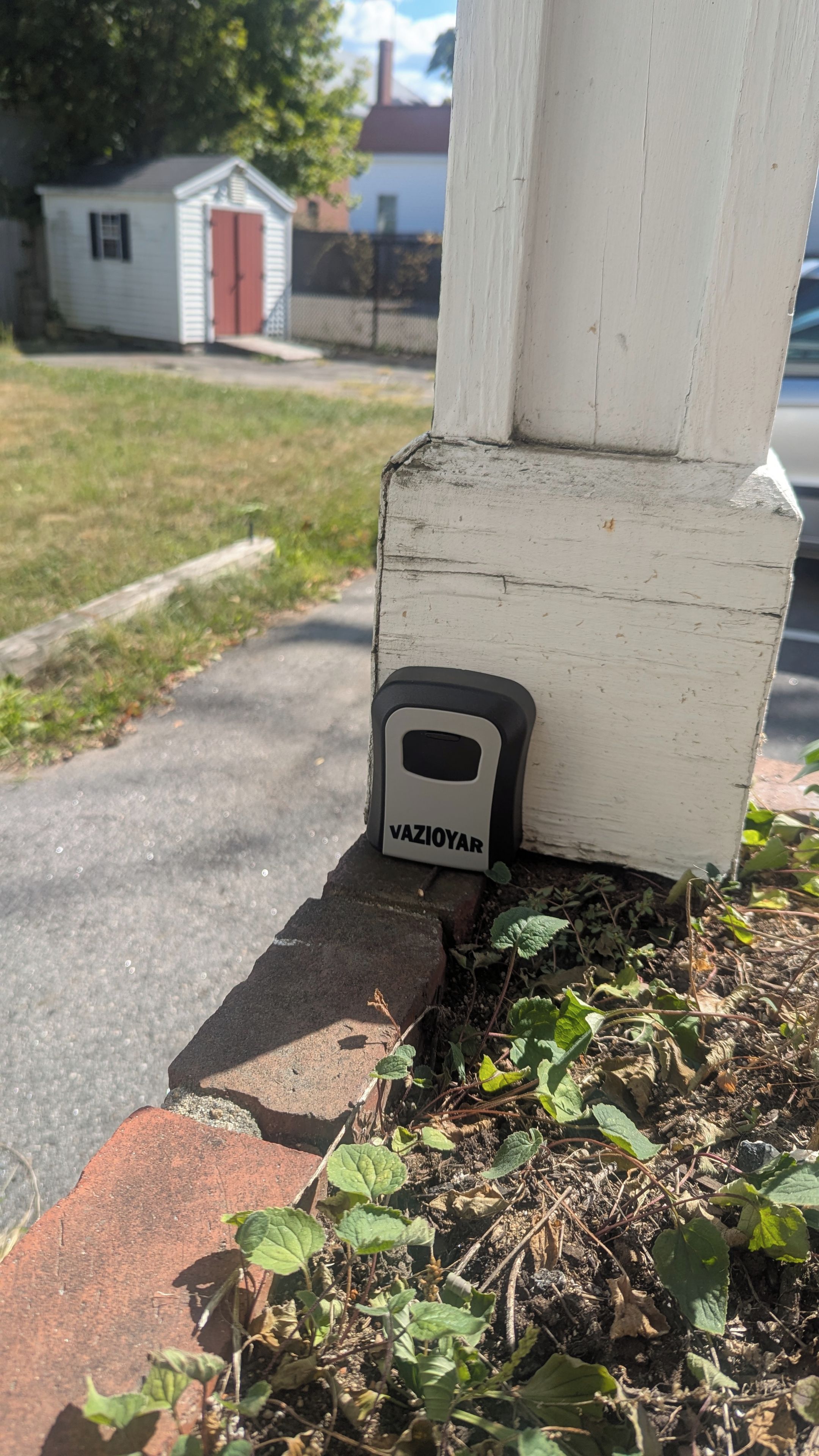 Un dispositif de sécurité portant la mention « VAZIOYAR » est fixé à un poteau blanc près d’une base en briques. On aperçoit, à l’arrière-plan, une zone herbeuse et un petit bâtiment.