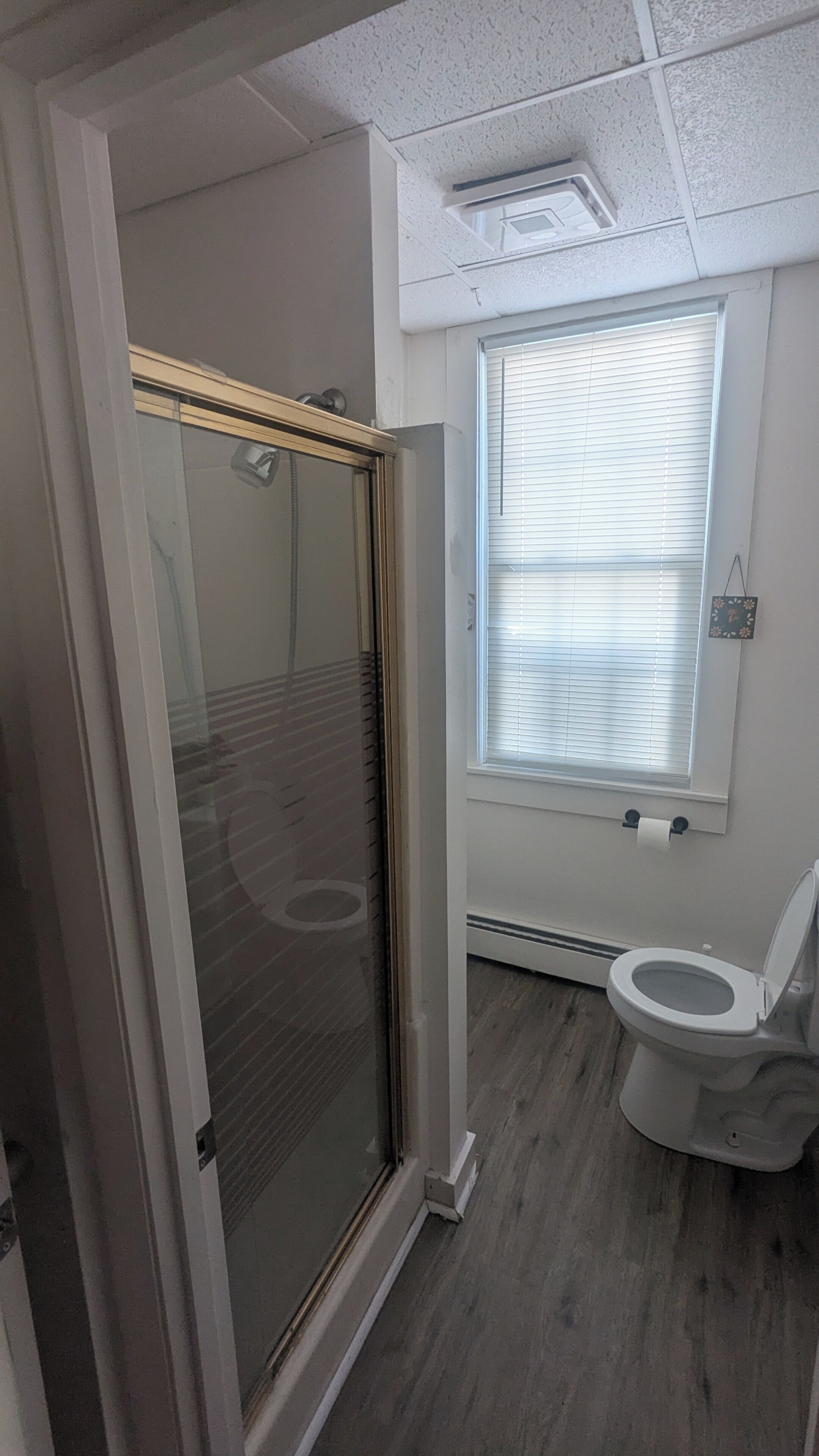 Une salle de bain comprenant des toilettes, une douche avec une porte en verre et une fenêtre avec des stores.