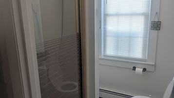 Une salle de bain comprenant des toilettes, une douche avec une porte en verre et une fenêtre avec des stores.