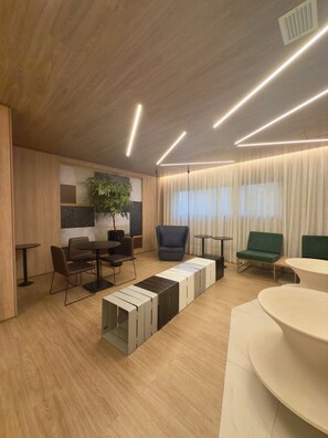 A modern interior with wooden flooring, a seating area with tables and chairs, a large plant, and a window with curtains. - Air Brooklin Residence (São Paulo)