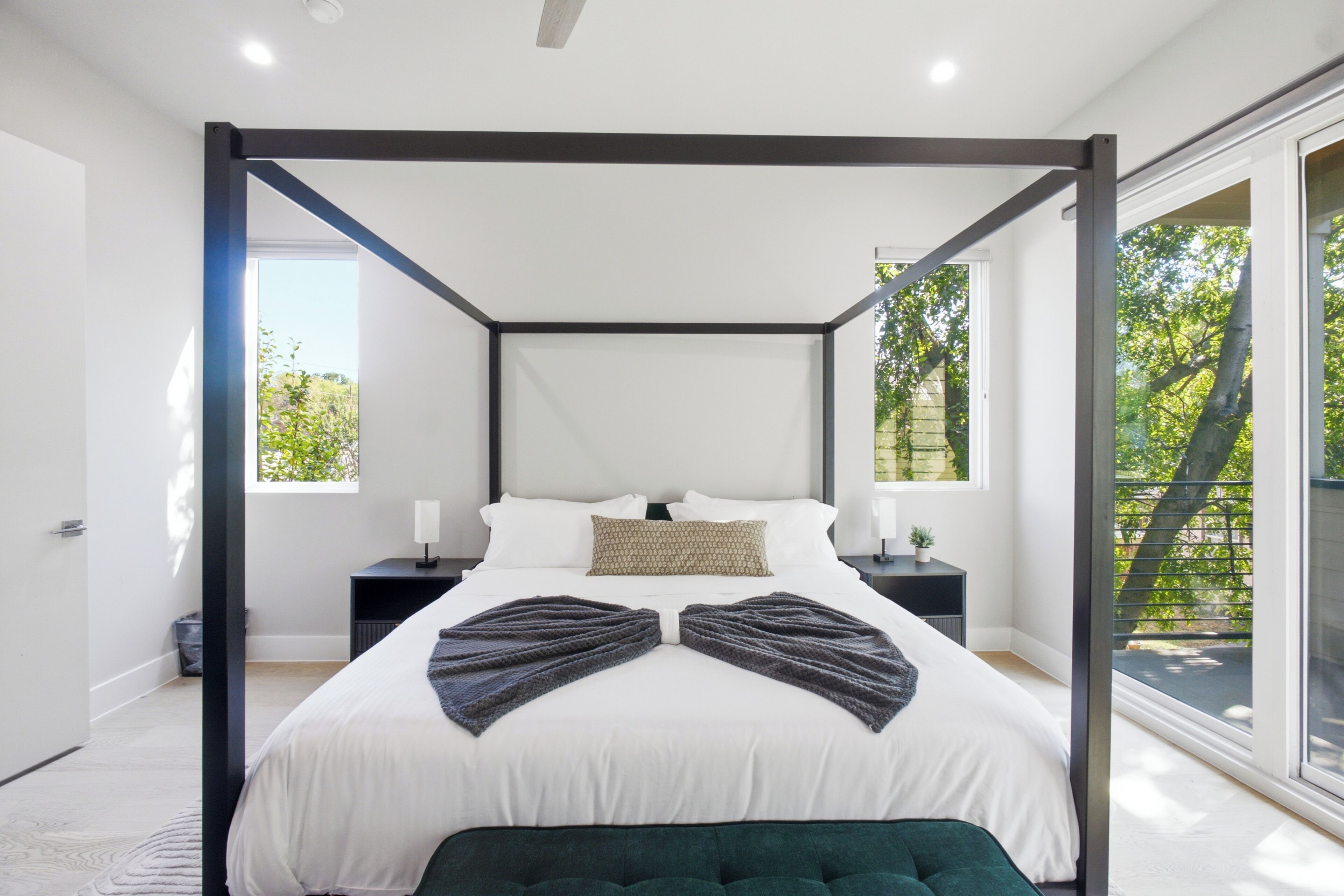 Quarto moderno com cama de dossel, mesa de cabeceira com abajur e vista para o verde através de amplas janelas.