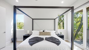 A modern bedroom with a four-poster bed, a bedside table with a lamp, and a view of greenery through large windows. - Dallas LUXE 4BR, 4 Kings, Hot Tub, Outdoor Theater (Dallas)