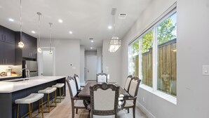 A modern kitchen with a central island, bar stools, and a dining area with a large table and chairs. - Dallas LUXE 4BR, 4 Kings, Hot Tub, Outdoor Theater (Dallas)