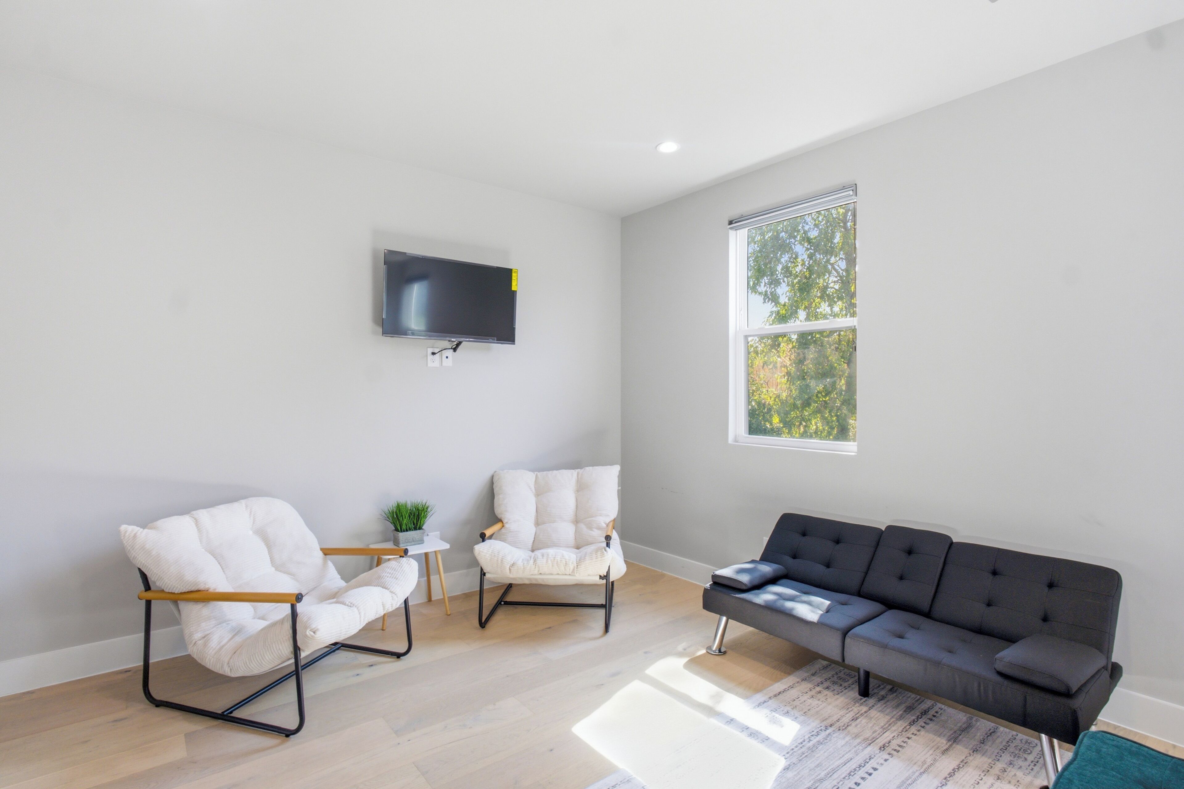 A living room with a flat-screen TV mounted on the wall, two white armchairs, a dark grey sofa, and a large window providing natural light.