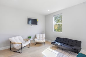 Ein Wohnzimmer mit einem wandmontierten Flachbildfernseher, zwei weißen Sesseln, einem dunkelgrauen Sofa und einem großen Fenster, das für natürliches Licht sorgt.
