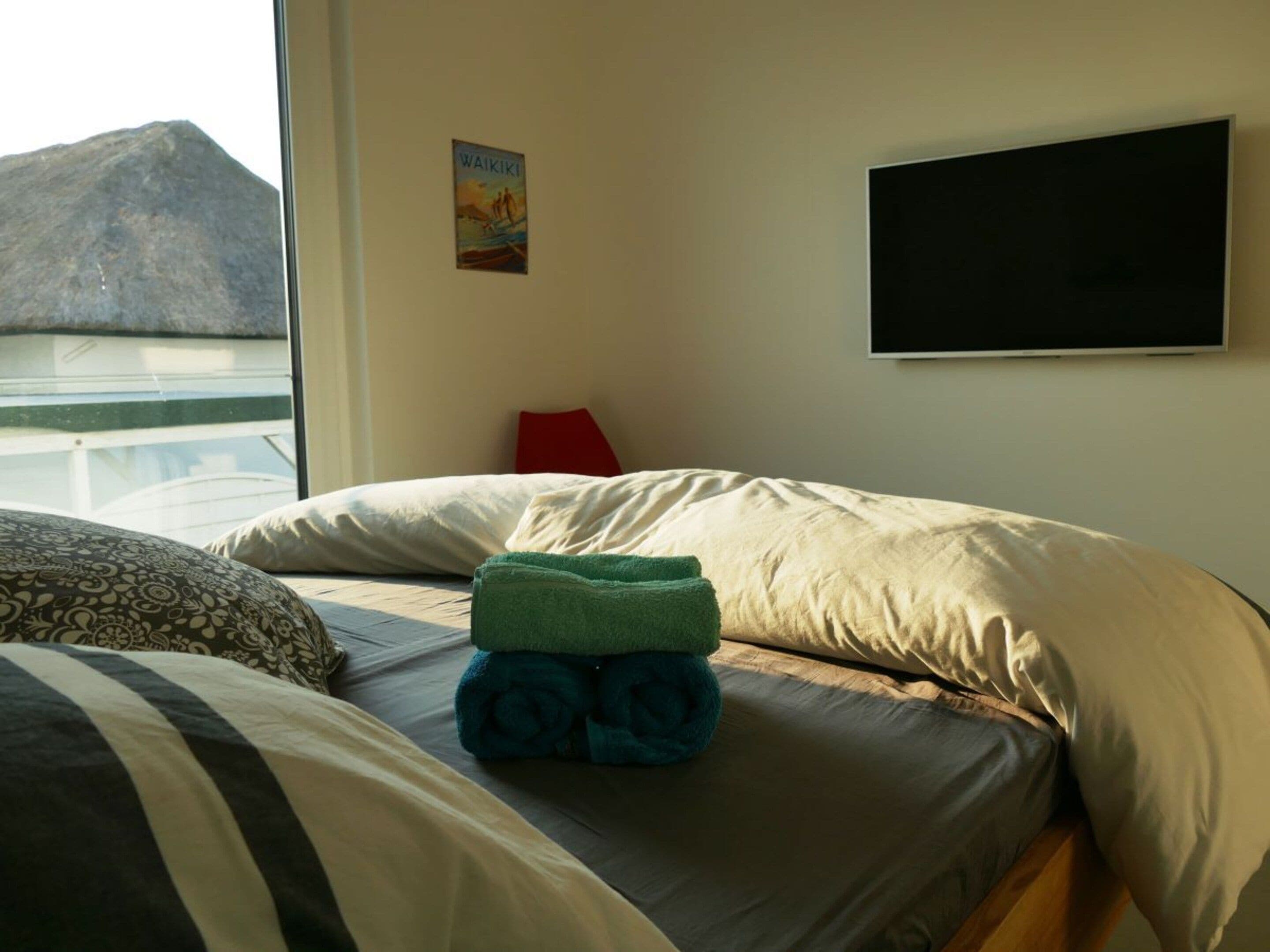 Quarto com cama, almofadas, toalha enrolada, pôster na parede e janela com vista para uma montanha.