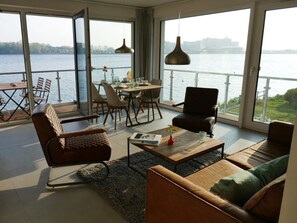 A spacious living area with a large window overlooking a body of water, furnished with a sofa, armchair, and a coffee table. - House Kiki, Apartment 2 "waikiki" (Heiligenhafen)