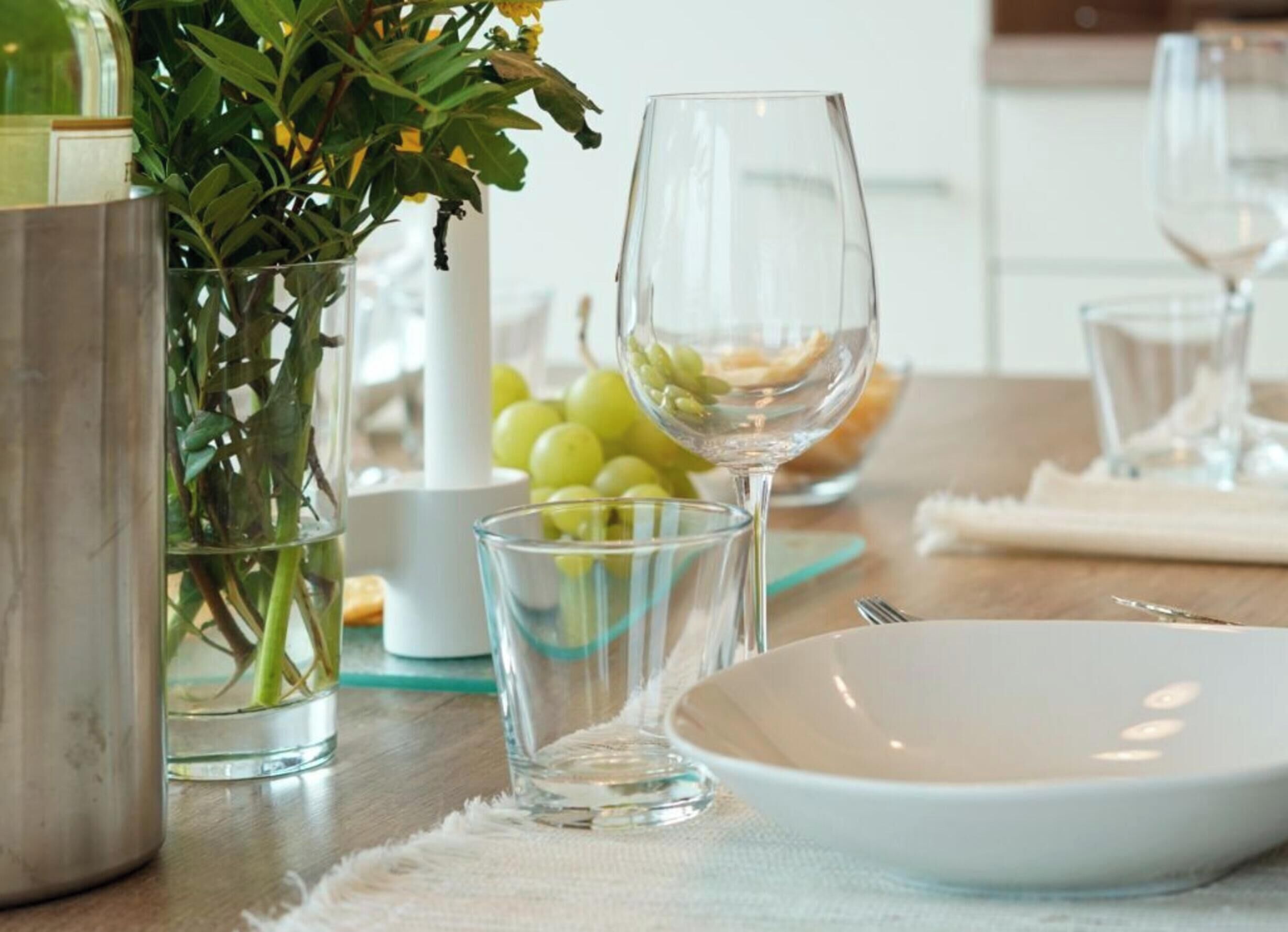 Ein Esstisch mit weißem Teller, einer Glasbowl und einem Glas Wasser. Grüne Trauben und ein klares Glas sind ebenfalls vorhanden.