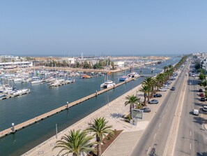 Eine Marina mit vertäuten Booten entlang einer Uferpromenade, eine Straße parallel zum Wasser und Palmen säumen den Gehweg.