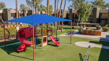 A playground with a red slide and climbing structures, surrounded by palm trees and a covered seating area.