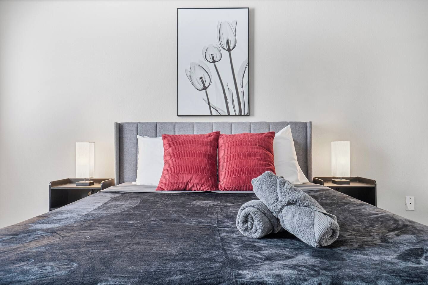 A modern bedroom with a grey headboard, a marble bedspread, two red pillows, a rolled-up towel, and bedside lamps.