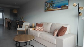 Ein modernes Wohnzimmer mit Sofa, Stühlen, einem Essbereich und einem Gemälde einer Küstenlandschaft.