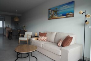Una sala de estar moderna con un sofá, sillas, un comedor y un cuadro con una escena costera.