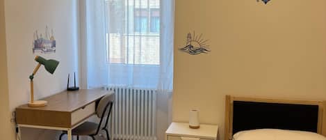 Une chambre à coucher avec un lit, un bureau, une chaise, un radiateur et une fenêtre avec des rideaux.