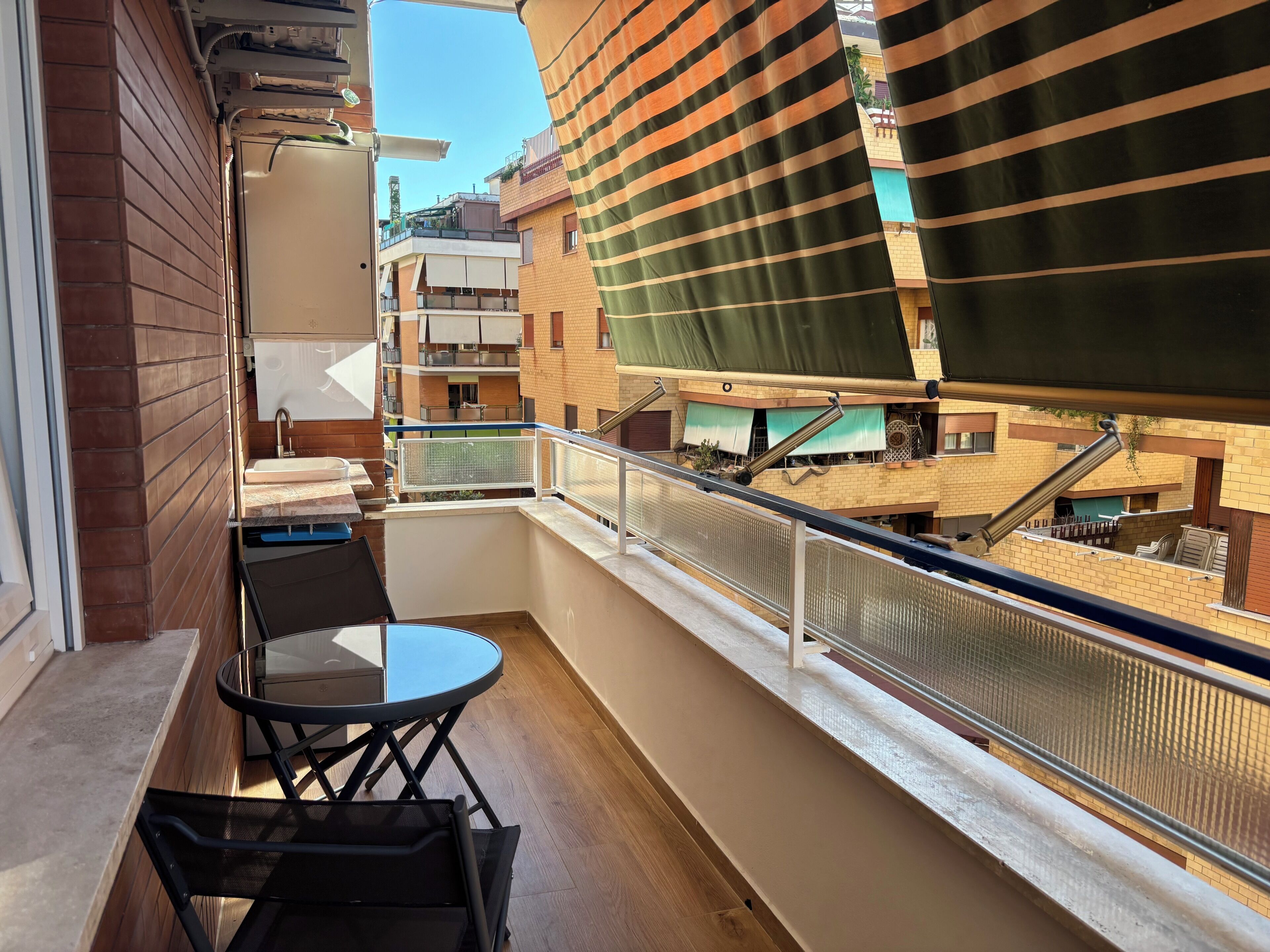 A balcony with a table and chairs, a view of adjacent buildings, and a metal railing.