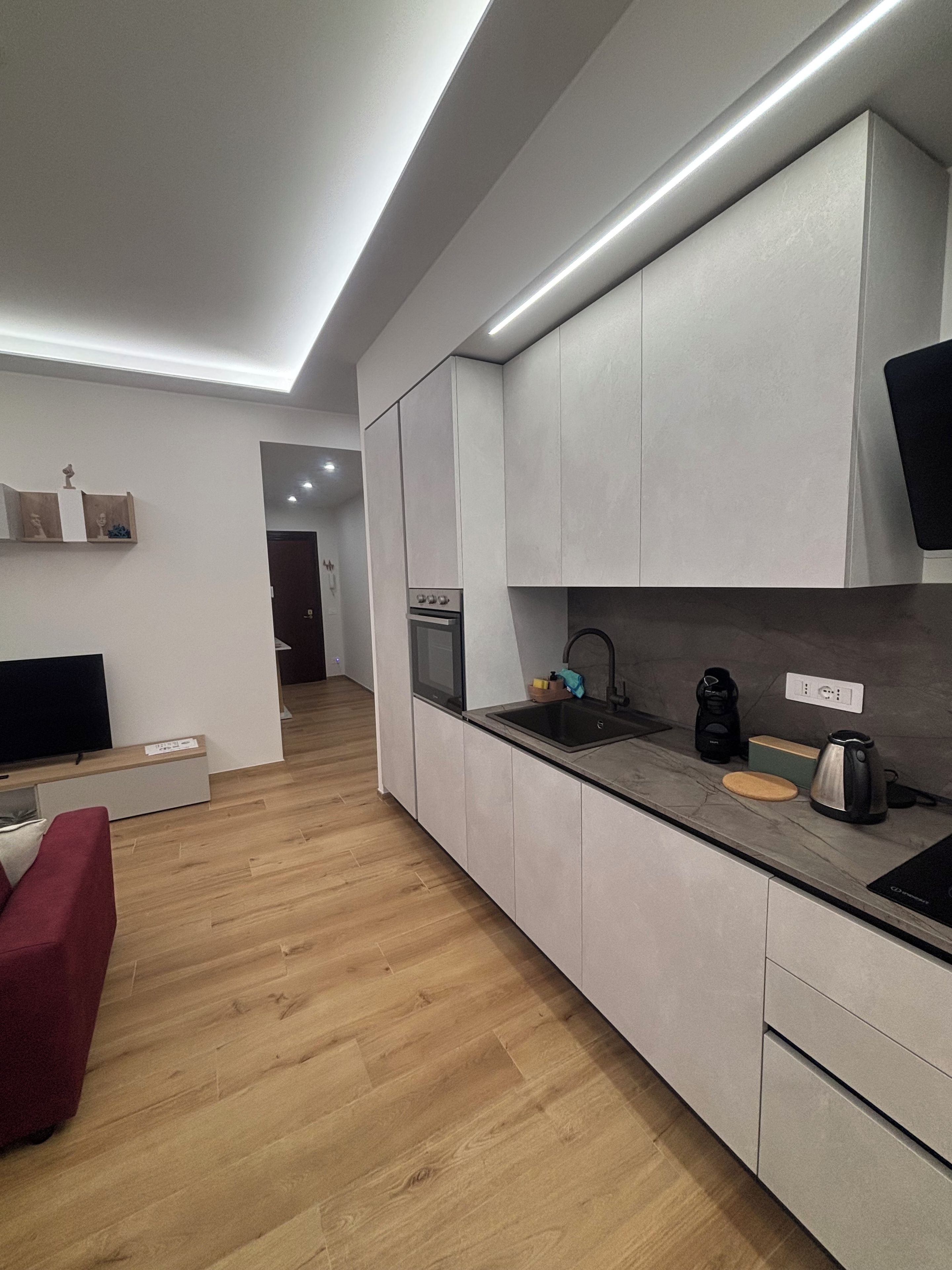 A modern kitchen with a built-in oven, a sink, and a coffee maker. There is a red couch and a flat-screen TV in the adjacent living area.