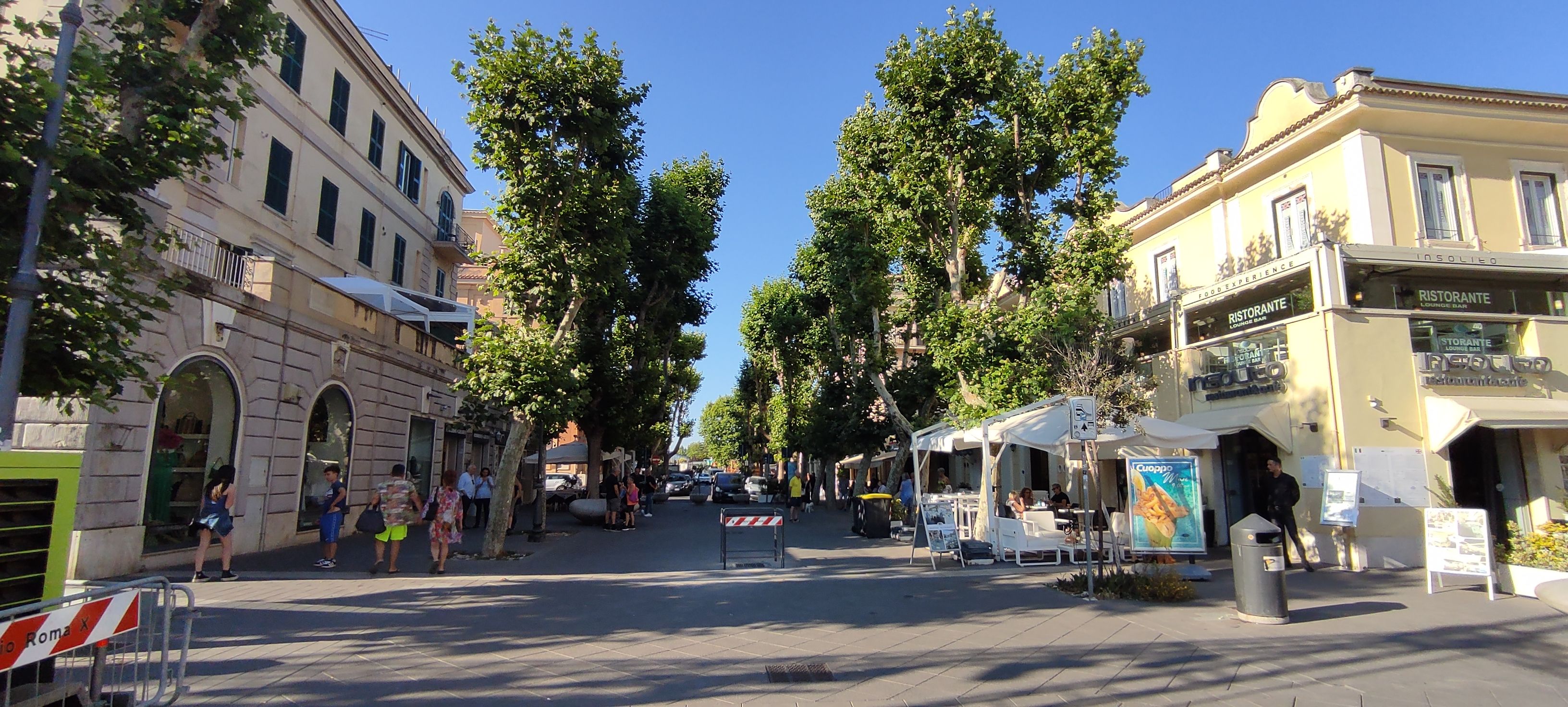 Una strada con tavolini all'aperto, alberi e edifici con insegne di ristoranti e bar.