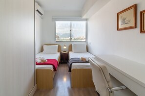 A compact hotel room with two single beds, a small table, a chair, a window with a view, and a wall-mounted air conditioning unit. - 2BR Apt Just 10 Mins From Barra da Tijuca Beach Vlm905 (Rio de Janeiro)