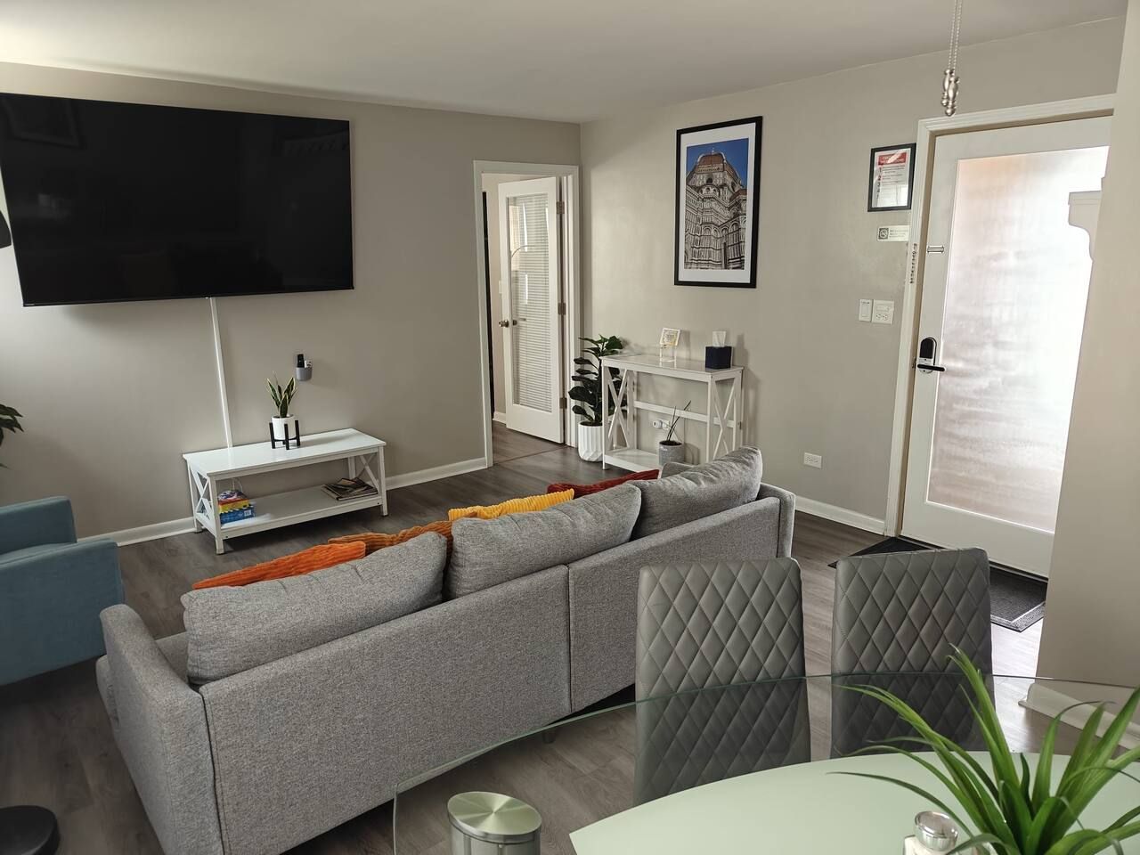 A modern living room with a grey sofa, a flat-screen TV, a white console table, and a glass dining table.