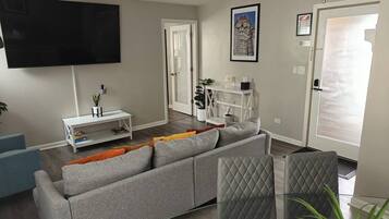 A modern living room with a grey sofa, a flat-screen TV, a white console table, and a glass dining table.
