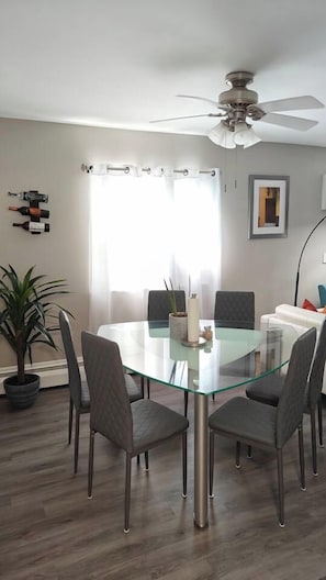 A dining area with a glass table, six chairs, a ceiling fan, a potted plant, and a window with curtains. - Greenwood Loft & Rooftop (Waukegan)