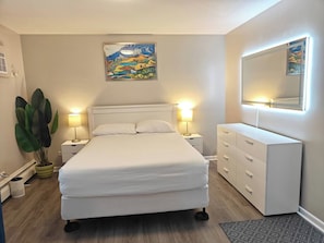 A bedroom with a bed, bedside lamps, a dresser, a large plant, and a painting on the wall. - Greenwood Loft & Rooftop (Waukegan)