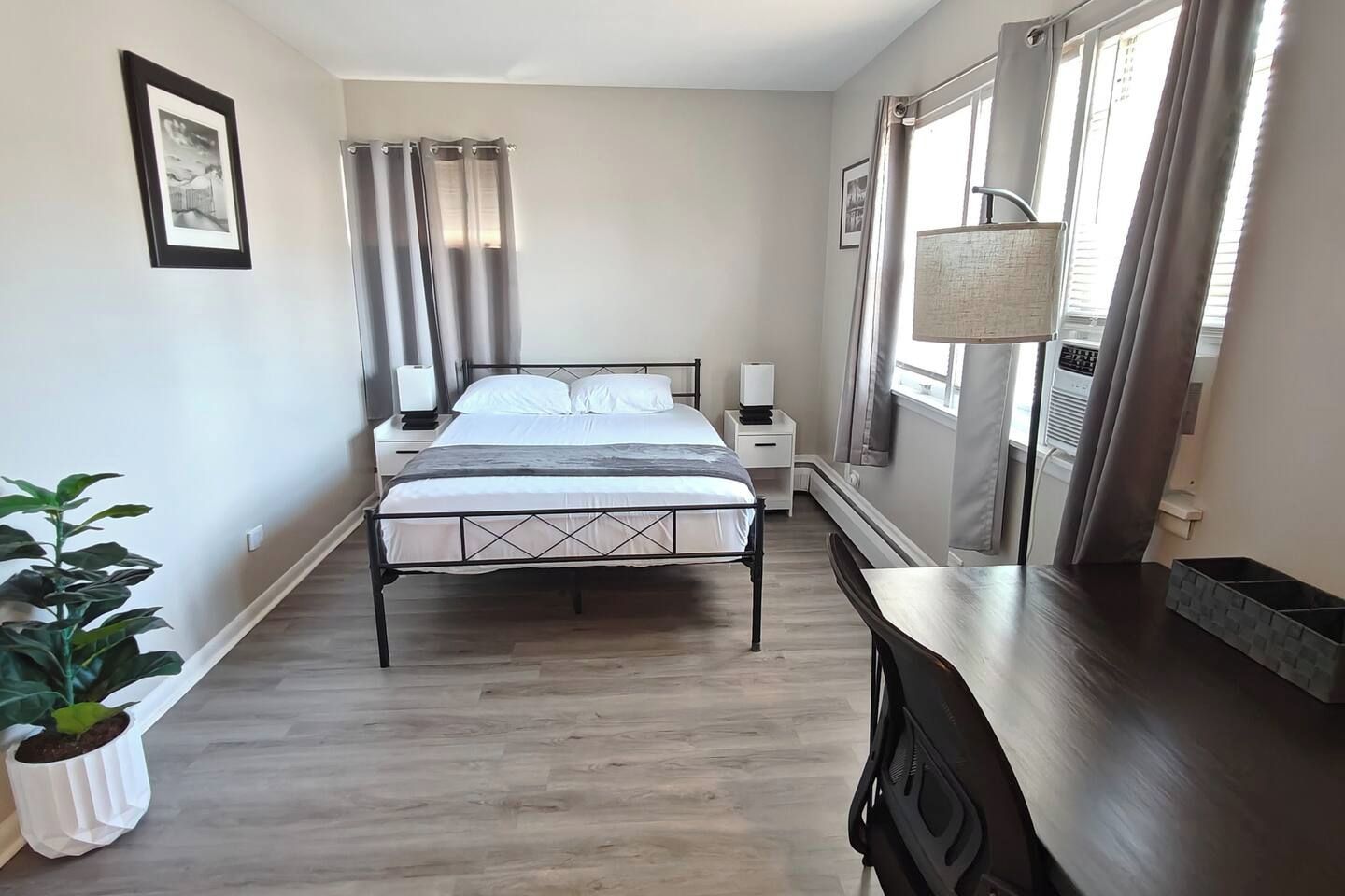 Habitación de hotel con cama, mesitas de noche, escritorio, silla, planta en maceta y ventana con cortinas.